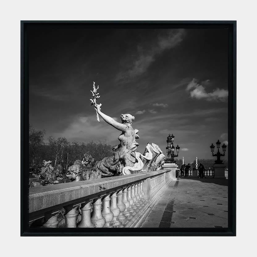 Oeuvre La Paix encadrée. Fine art photographie en noir et blanc d'un paysage urbain de Bordeaux. Sur la place des Quinconces aux monuments aux girondins, nous voyons une femme au centre de l'image. Elle brandit conquérante un rameau d'olivier, symbole de paix, titre de l'oeuvre. Le regard est accompagné jusqu'au sujet par une belle profondeur de champs venant en diagonale inférieure gauche. C'est une rampe d'époque en colonne qui pierre qui dynamise l'image tout en servant le sujet photographique. Lumineuse et contrastée, l'image semble prendre vie grâce aux nuages, comme en mouvement
