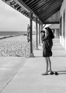 Photographie noir et blanc d'Anne-Laure Boisseau en train de photographier l'océan sous le porche d'une terrasse. Cette image représente bien l'artiste lors de sa prise de vue, décontractée en pleine nature, profitant de l'instant présent.