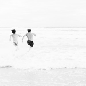 Photographie d'art en noir et blanc, représentant deux hommes faisant la course pour rentrer dans l'océan atlantique. Vu de dos, le mouvement figé de leur corps pressés rappelle le cartoon du Coyote. Les embruns de l'eau se soulèvent derrière leur passage.