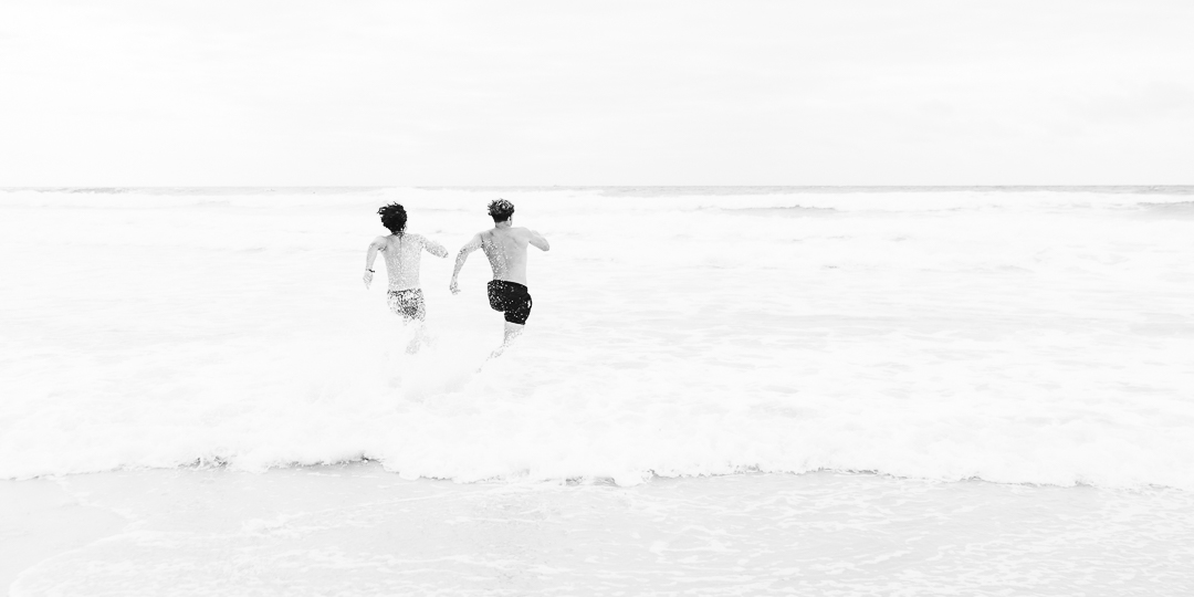 Photographie d'art en noir et blanc, représentant deux hommes faisant la course pour rentrer dans l'océan atlantique. Vu de dos, le mouvement figé de leur corps pressés rappelle le cartoon du Coyote. Les embruns de l'eau se soulèvent derrière leur passage.
