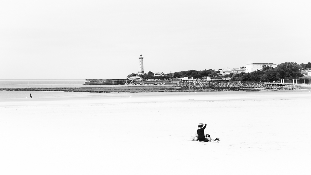 Photographie d'art en noir et blanc d'une femme profitant d'un instant privilégier sur la plage de St-Georges-de-Didonne avec son fils. Ce cliche, comme un paysage dessiné avec son magnifique phare, raconte une histoire apaisée grâce à la présence du personnage. Elle nous projette davantage dans l'emotion, le sentiment que l'on ressent en bord d'ocean. Et l'on s'evade quelques instants...