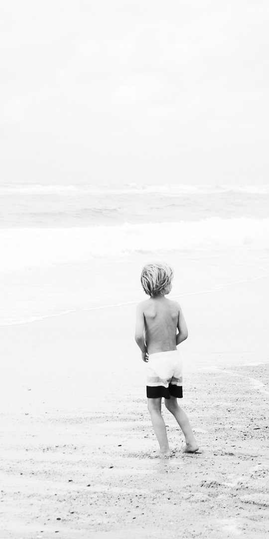 Jeune enfant blond, de dos, seul sur la plage et face à l’océan. Il regarde vers la droite : appel silencieux de l’océan, dans la liberté de l’enfance. Photo en noir et blanc.