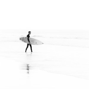 Photographie en noir et blanc d’un surfeur marchant seul sur le rivage avec sa planche de surf, capturé dans un paysage minimaliste baigné de lumière, créant une sensation de liberté et de sérénité.
