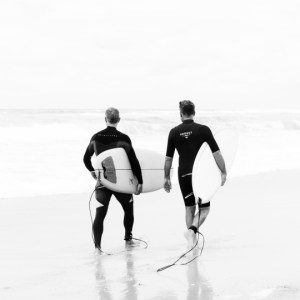 Deux surfeurs marchant côte à côte, planches sous le bras, regardant l'océan. Leurs silhouettes presque identiques traduisent une complicité subtile et une communion avec la mer, capturées dans une scène épurée en noir et blanc.
