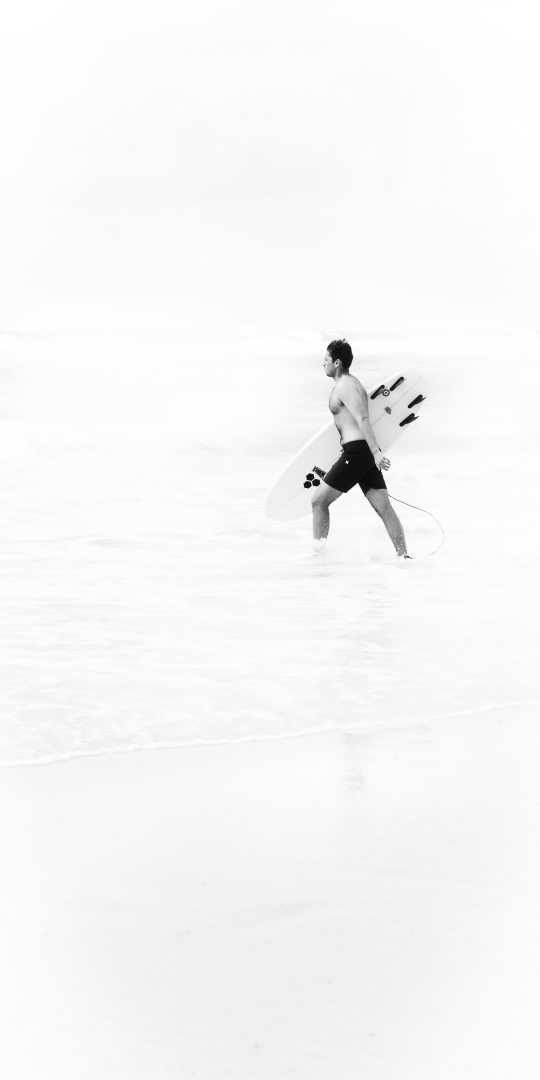Un surfeur avance avec confiance, planche sous le bras, vers l'océan. La photographie en noir et blanc met en avant sa silhouette expressive, accentuée par un jeu de lumière et de contrastes qui pousse l’essentiel dans l’épure, évoquant à la fois l'excitation et la sérénité.