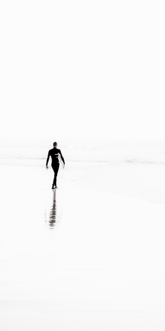Photographie en noir et blanc d’un surfeur marchant avec élégance vers l’horizon, sa silhouette et sa réflexion créant une scène minimaliste et poétique.