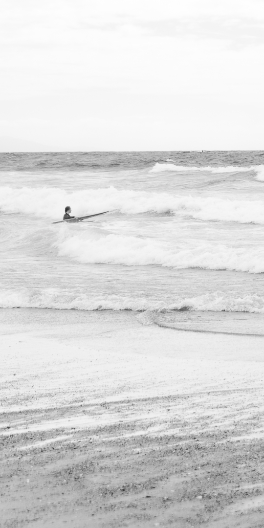 Une surfeuse avance dans l’océan, affrontant les premières vagues avec calme et humilité, capturée en noir et blanc dans un moment de connexion et de respect face à la mer.