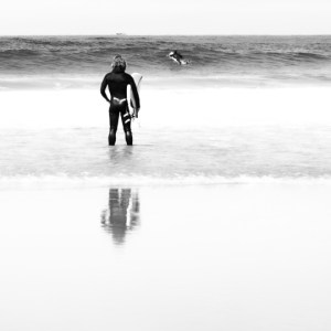 Photographie en noir et blanc d’un surfeur immobile face à l’océan, absorbé par l’instant. Sa posture calme contraste avec la puissance latente de sa silhouette dynamique, exprimant une connexion intime avec l'eau et une intensité visuelle captivante.