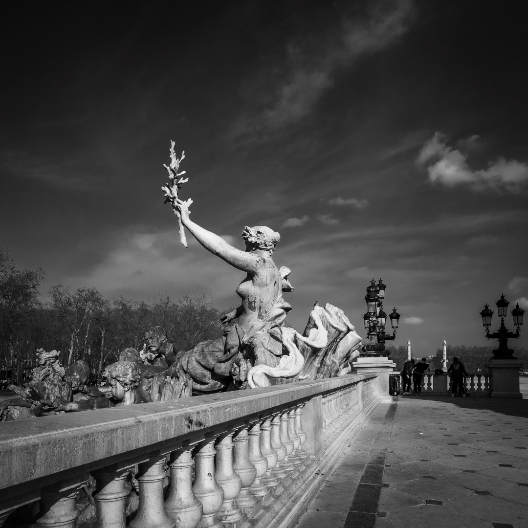 Fine art photographie en noir et blanc d'un paysage urbain de Bordeaux. Sur la place des Quinconces aux monuments aux girondins, nous voyons une femme au centre de l'image. Elle brandit conquérante un rameau d'olivier, symbole de paix, titre de l'oeuvre. Le regard est accompagné jusqu'au sujet par une belle profondeur de champs venant en diagonale inférieure gauche. C'est une rampe d'époque en colonne qui pierre qui dynamise l'image tout en servant le sujet photographique. Lumineuse et contrastée, l'image semble prendre vie grâce aux nuages, comme en mouvement